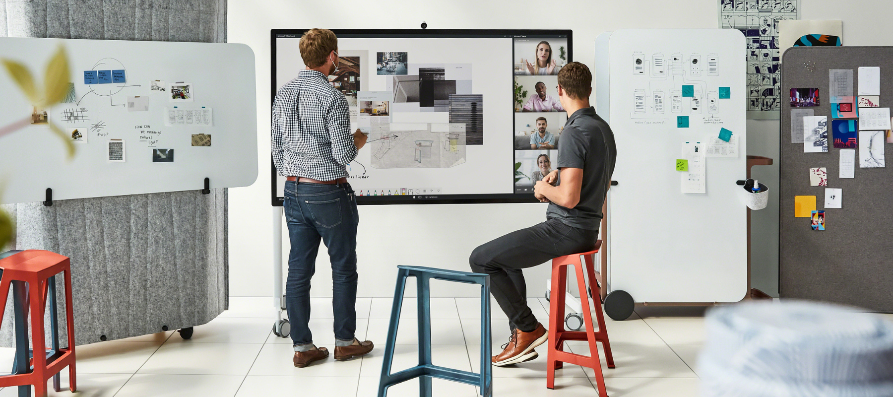 two people in office sitting on stools having a virtual meeting with other coworkers on a screen mounted on the wall